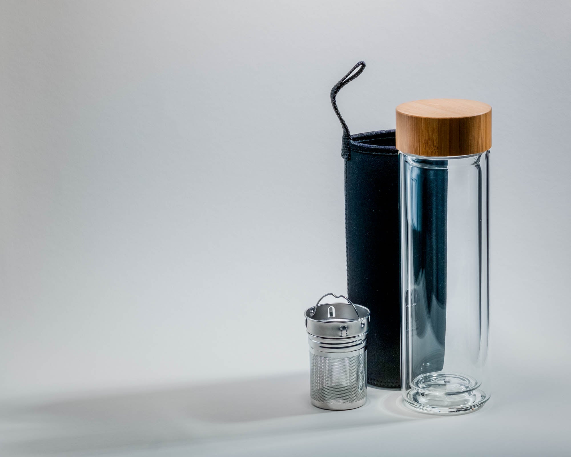 Eine elegante, transparente Glasflasche mit einem Bambusdeckel steht neben einer schwarzen Neoprenhülle mit Trageschlaufe und einem silbernen Teesieb. Der helle Hintergrund und die weichen Schatten betonen das minimalistische Design.