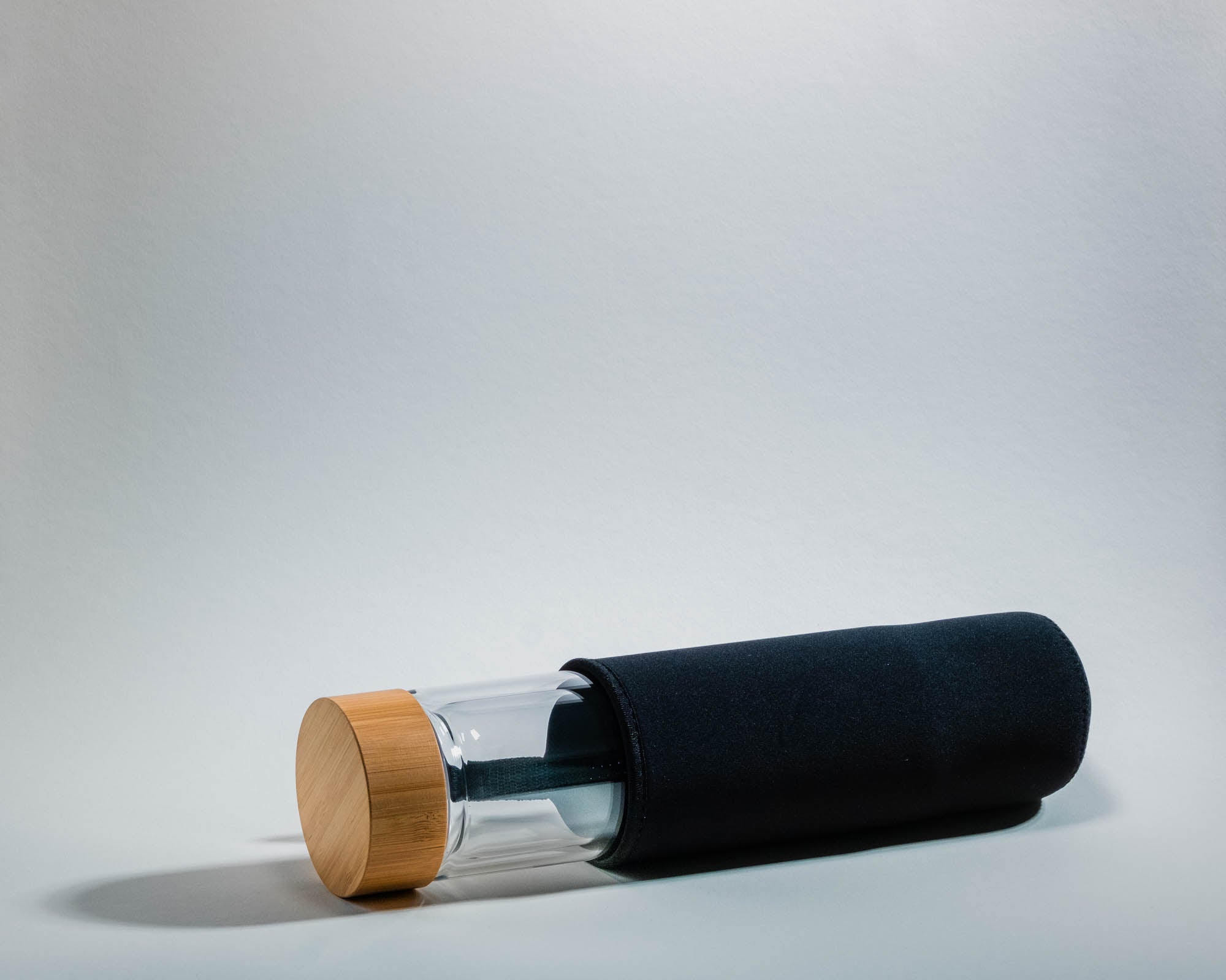 Eine liegende Glasflasche mit einem Bambusdeckel, teilweise in einer schwarzen Neoprenhülle. Die Flasche wirft einen weichen Schatten auf den hellen Hintergrund. Die Hülle schützt die Flasche und hat eine sichtbare Lasche für einfaches Herausnehmen.