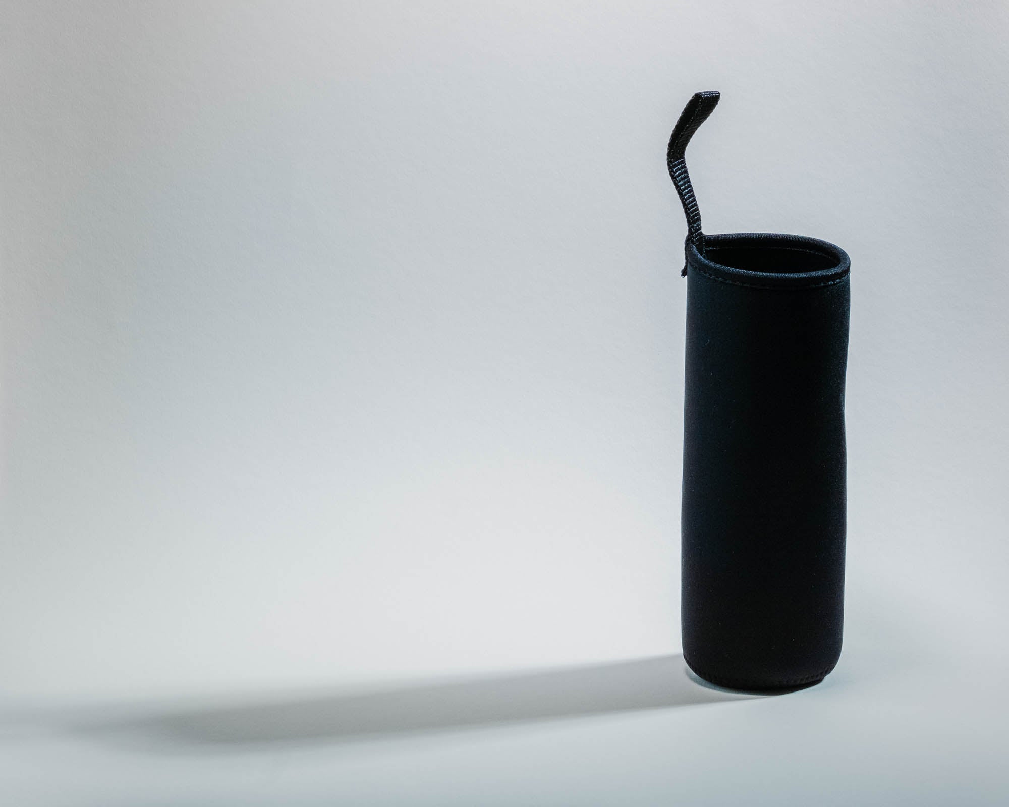 Eine schwarze, zylindrische Neoprenhülle mit einer kleinen, nach oben ragenden Schlaufe, stehend vor einem hellen Hintergrund. Die minimalistische und funktionale Gestaltung deutet auf eine Schutzhülle für eine Trinkflasche hin.
