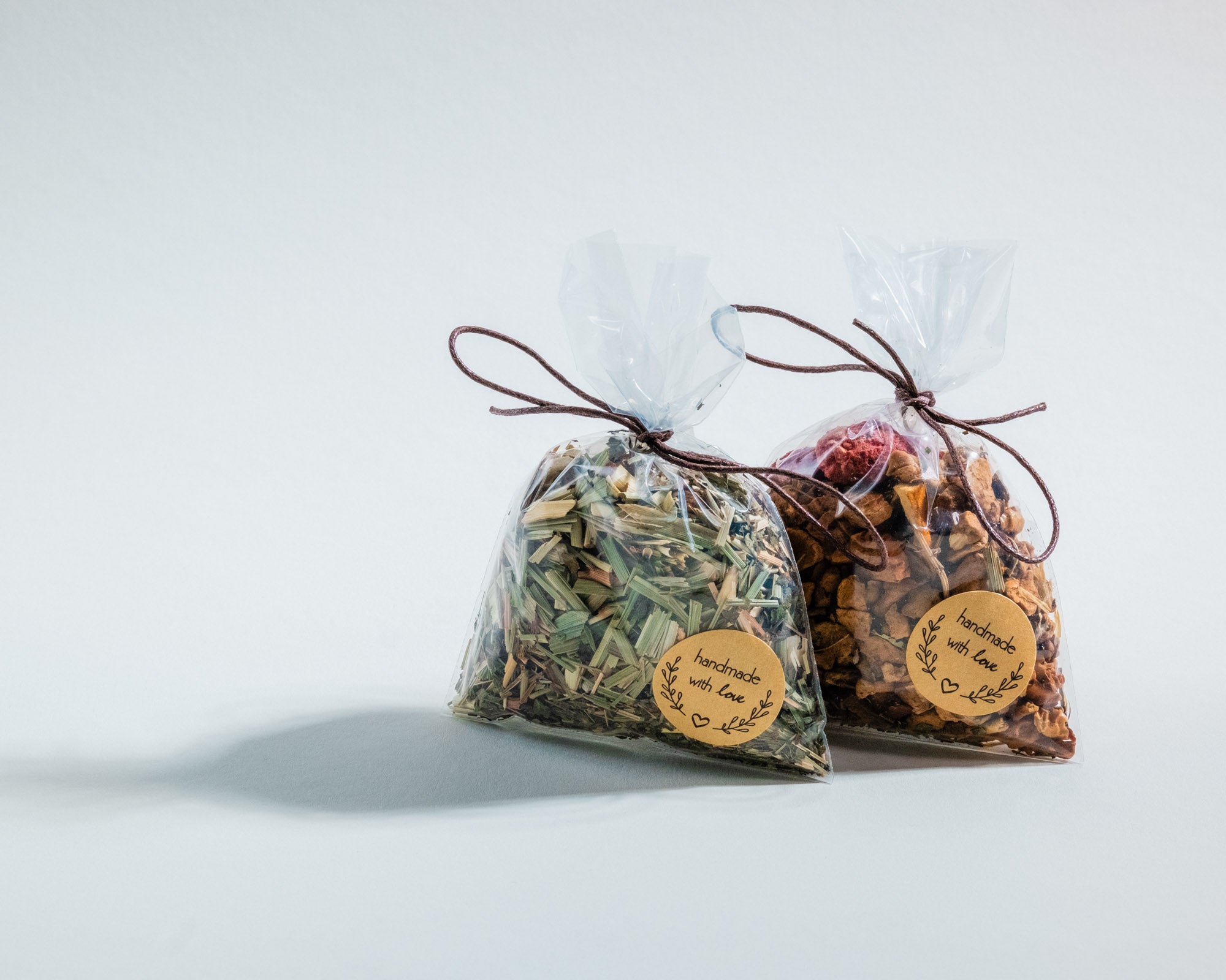 Zwei durchsichtige Geschenkbeutel mit Tee, einer mit getrockneten Kräutern und der andere mit Fruchtstücken. Beide sind mit einer braunen Kordel verschlossen und tragen ein rundes Etikett mit der Aufschrift „handmade with love“ sowie einem dekorativen Blätterkranz. Sie stehen auf einer weißen, neutralen Oberfläche und werfen weiche Schatten.