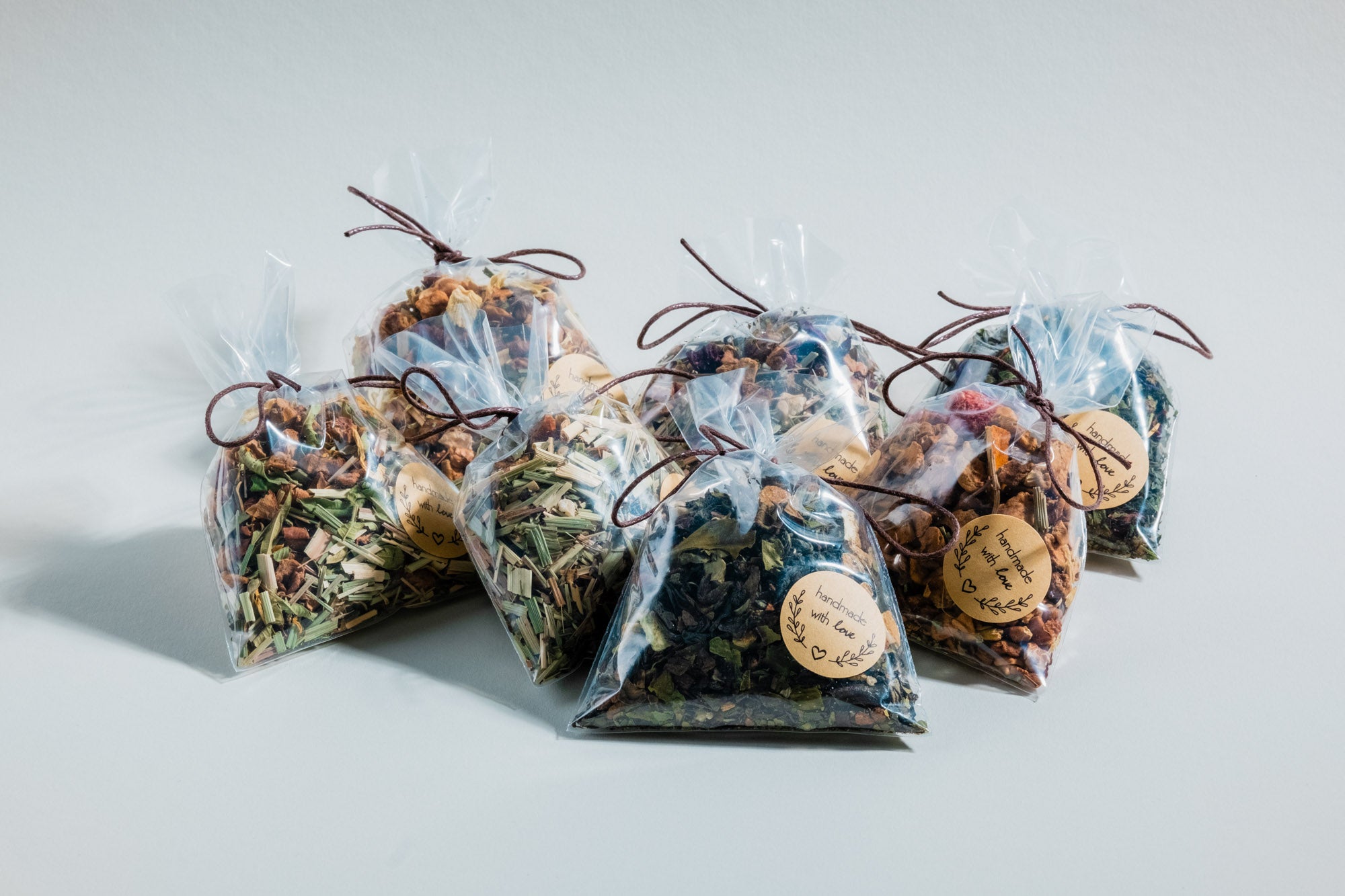 Sieben durchsichtige Geschenkbeutel mit verschiedenen Teemischungen, darunter Kräuter-, Frucht- und Blütentees. Jeder Beutel ist mit einer braunen Kordel verschlossen und trägt ein rundes Etikett mit der Aufschrift „handmade with love“ und einem dekorativen Blätterkranz. Sie sind auf einer neutralen, hellen Oberfläche arrangiert und werfen sanfte Schatten.