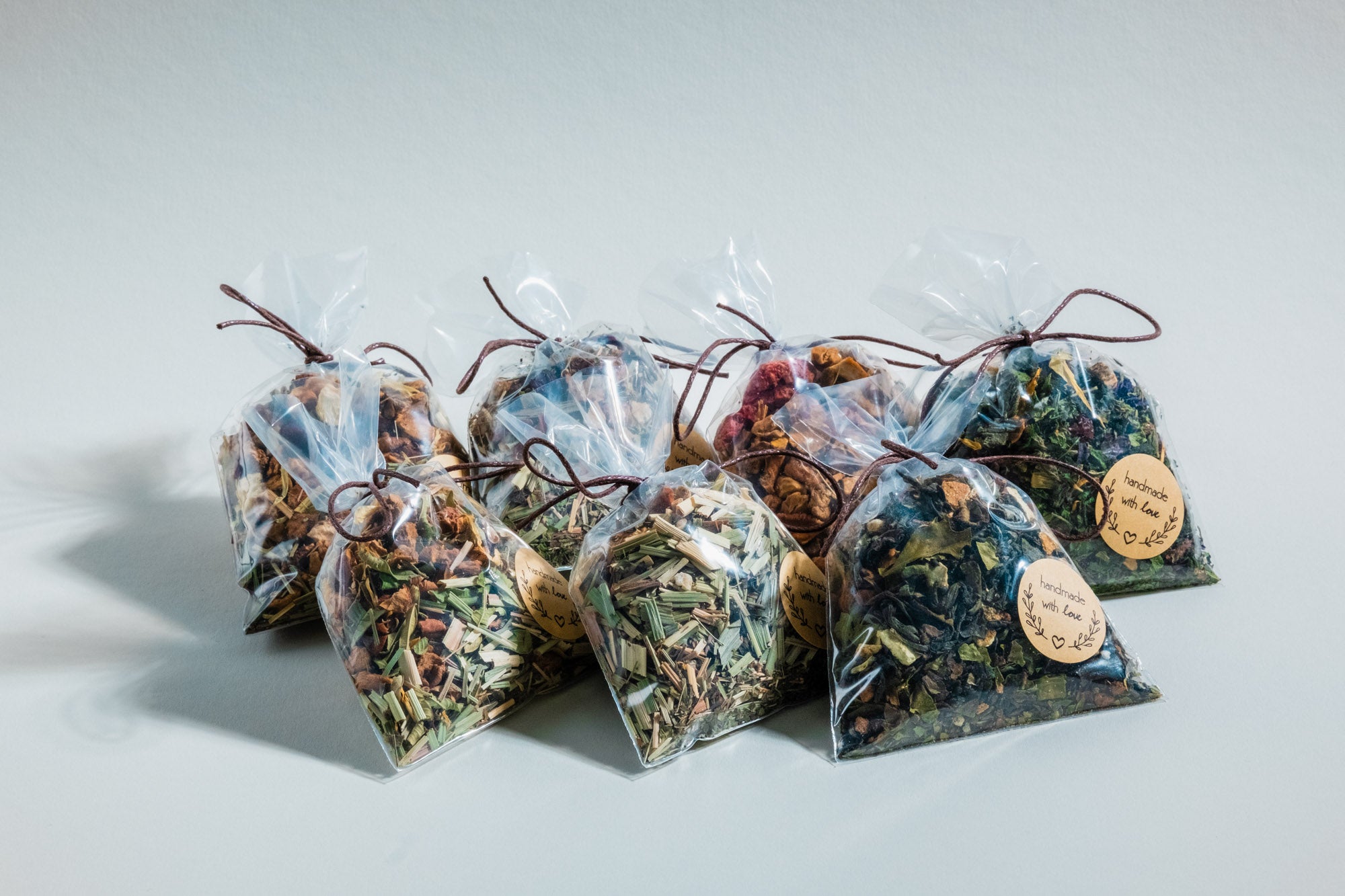 Sieben durchsichtige Geschenkbeutel mit verschiedenen Teemischungen, darunter Kräuter-, Frucht- und Blütentees. Jeder Beutel ist mit einer braunen Kordel verschlossen und trägt ein rundes Etikett mit der Aufschrift „handmade with love“ und einem dekorativen Blätterkranz. Sie sind auf einer neutralen, hellen Oberfläche arrangiert und werfen sanfte Schatten.