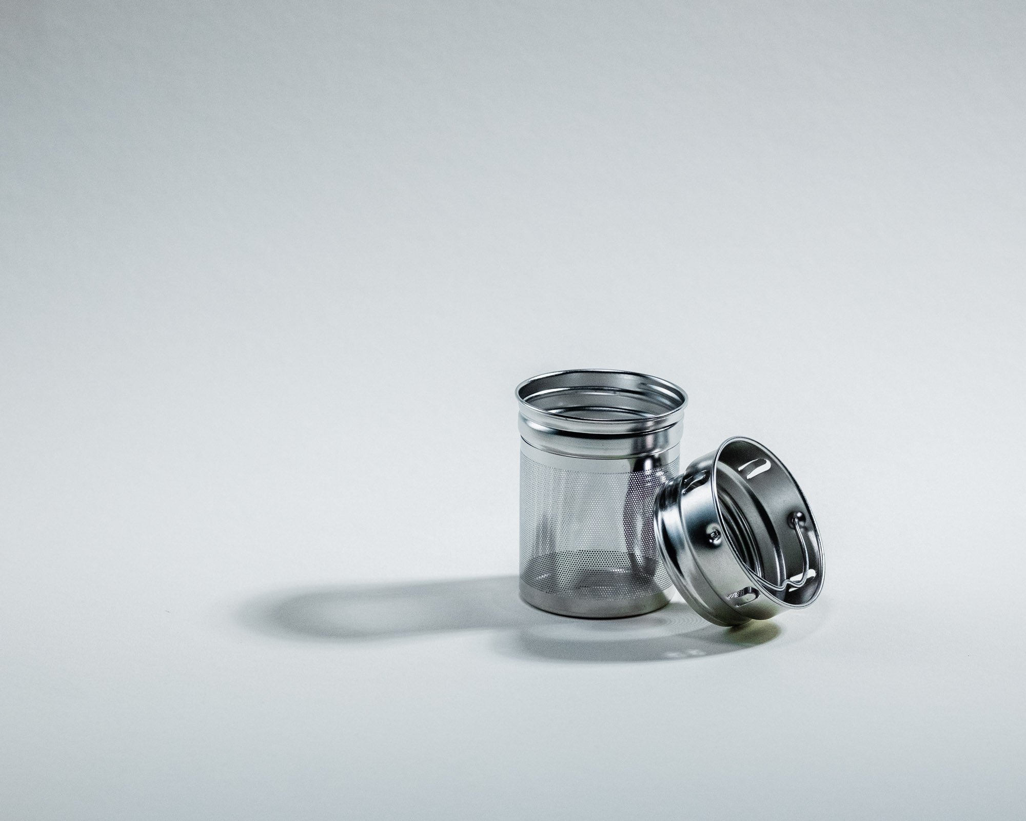 Ein zweiteiliges Teesieb aus Edelstahl, bestehend aus einem feinmaschigen Behälter und einem abgenommenen Deckel. Der Deckel liegt schräg neben dem Sieb auf einer weissen, neutralen Oberfläche, beide werfen leichte Schatten.