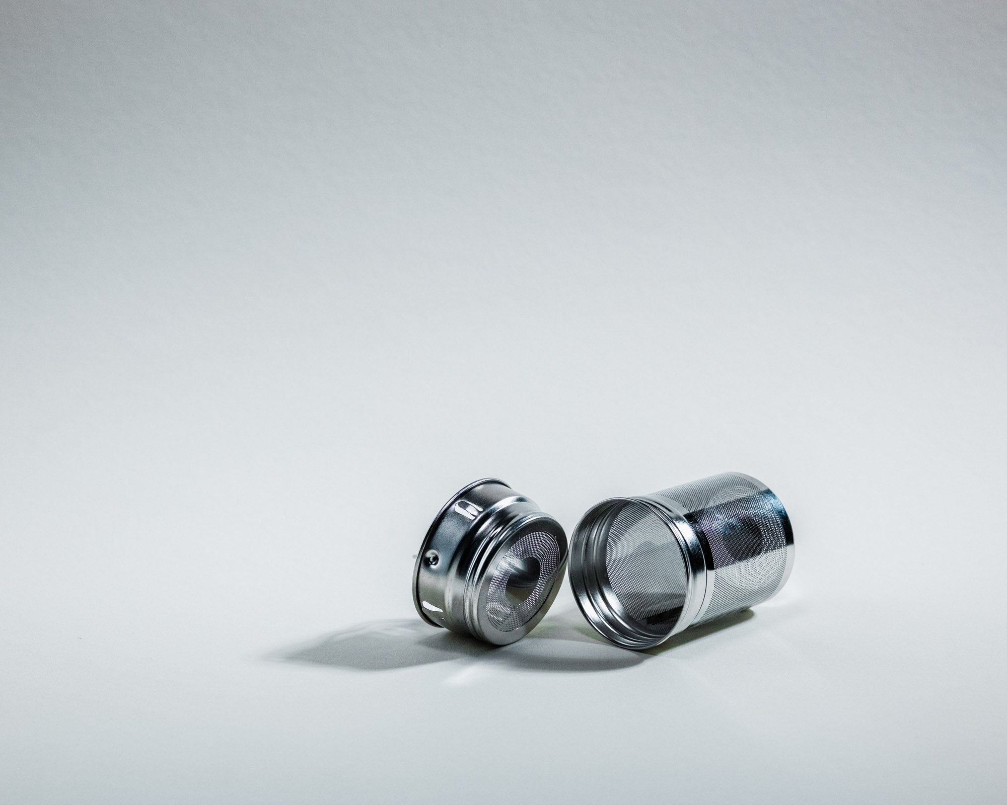 Ein auseinandergebautes Teesieb aus Edelstahl, bestehend aus zwei Teilen. Der feinmaschige Behälter liegt rechts, während der Deckel links daneben auf einer weissen, neutralen Oberfläche liegt. Beide Teile werfen leichte Schatten.
