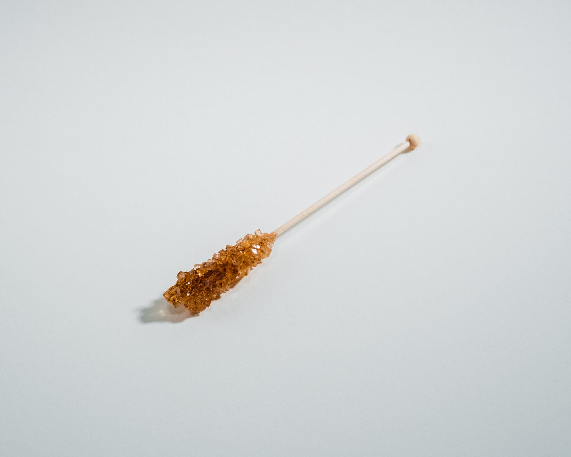 Ein einzelner brauner Kandiszucker-Stick mit großen, glänzenden Zuckerkristallen auf einem hellen Hintergrund. Der Holzstab hat ein kleines rundes Ende und wirft einen sanften Schatten.