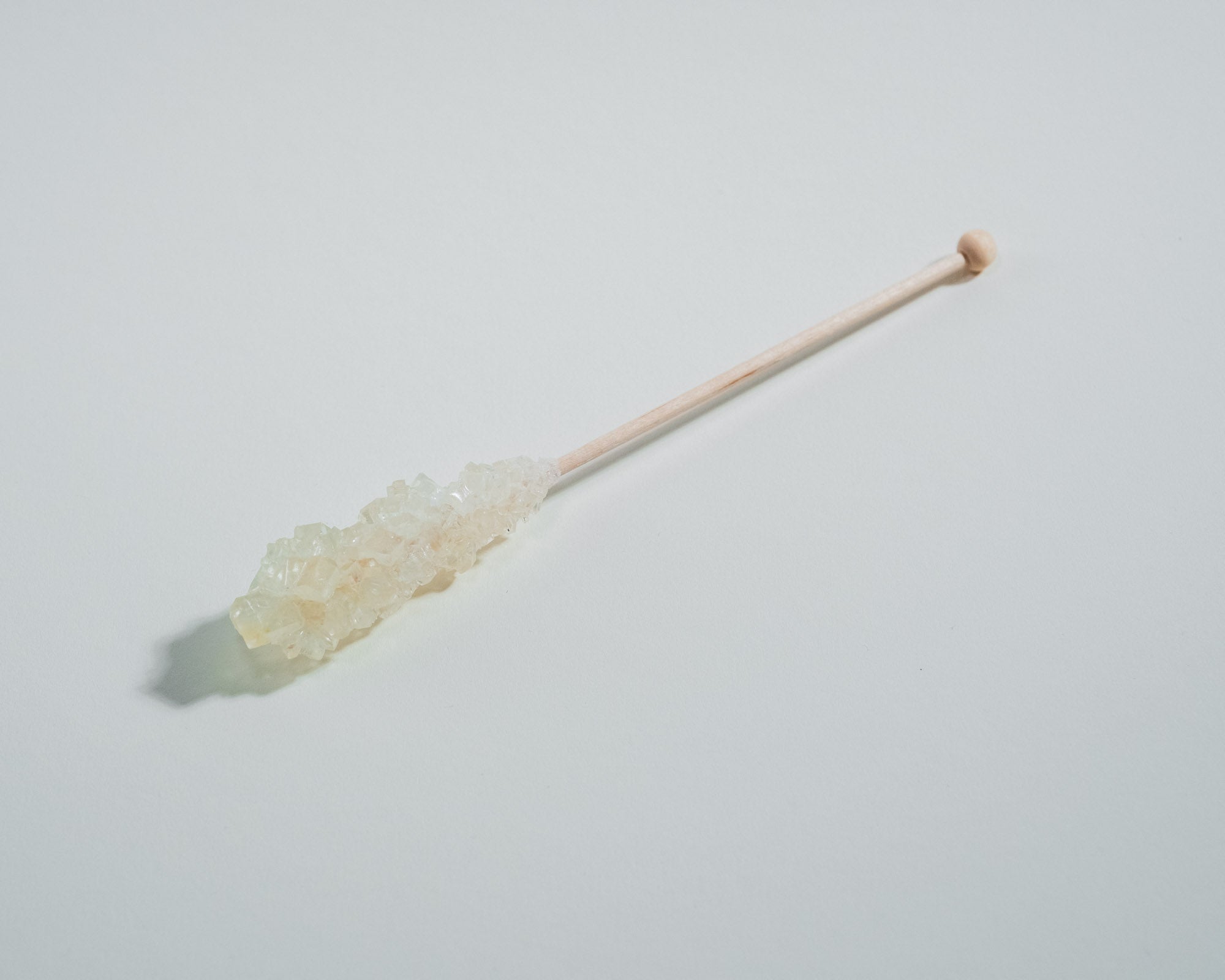 Ein einzelner weißer Kandiszucker-Stick mit großen, klaren Zuckerkristallen auf einem hellen Hintergrund. Der Holzstab hat ein rundes Ende und wirft einen sanften Schatten.
