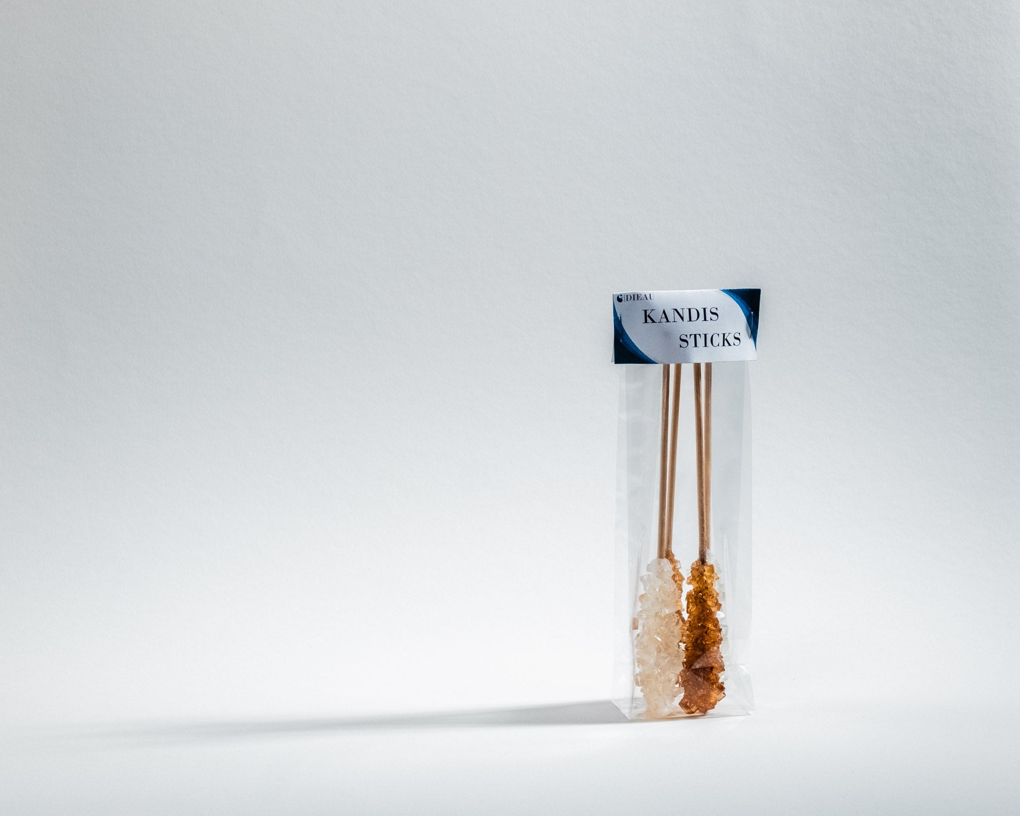 Eine transparente Verpackung mit vier Kandiszucker-Sticks, zwei weiße und zwei braune, auf einem hellen Hintergrund. Die Verpackung hat ein Etikett mit der Aufschrift 'KANDIS STICKS' und einem blauen Designelement.